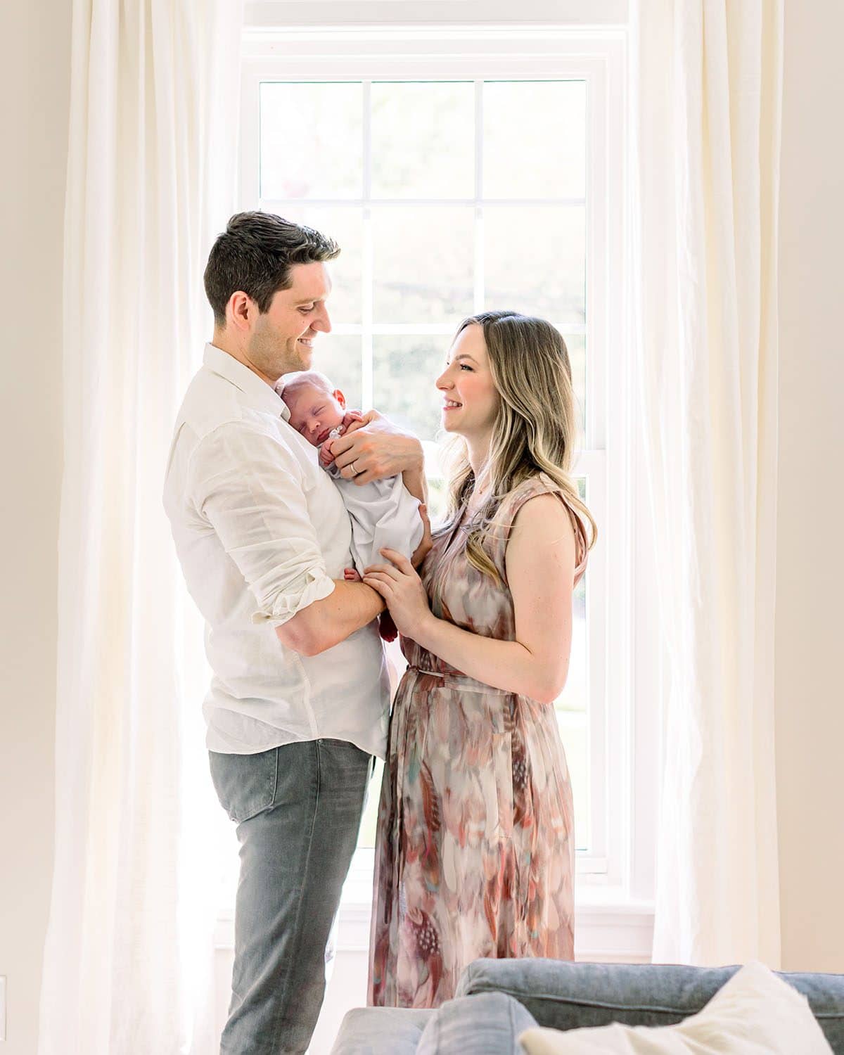 Parents standing by window holding baby by Miami Newborn Photographer