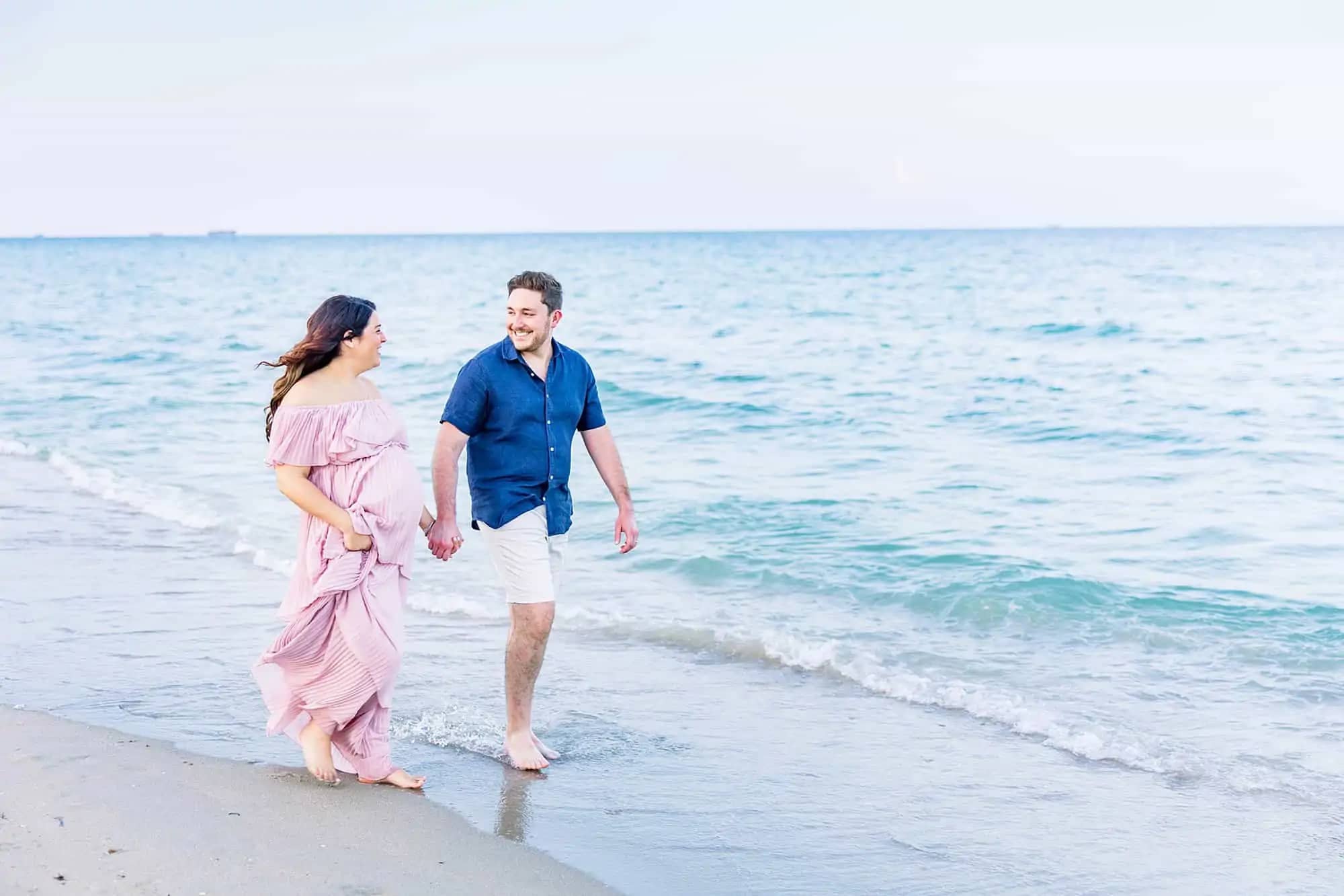 Couple walking holding hands on beach, pregnant woman is wearing pink ruffled tiered flowy dress, man is wearing dark blue button down and khaki shorts - What to Wear for Beach Maternity Photos | Maternity Photographer Miami